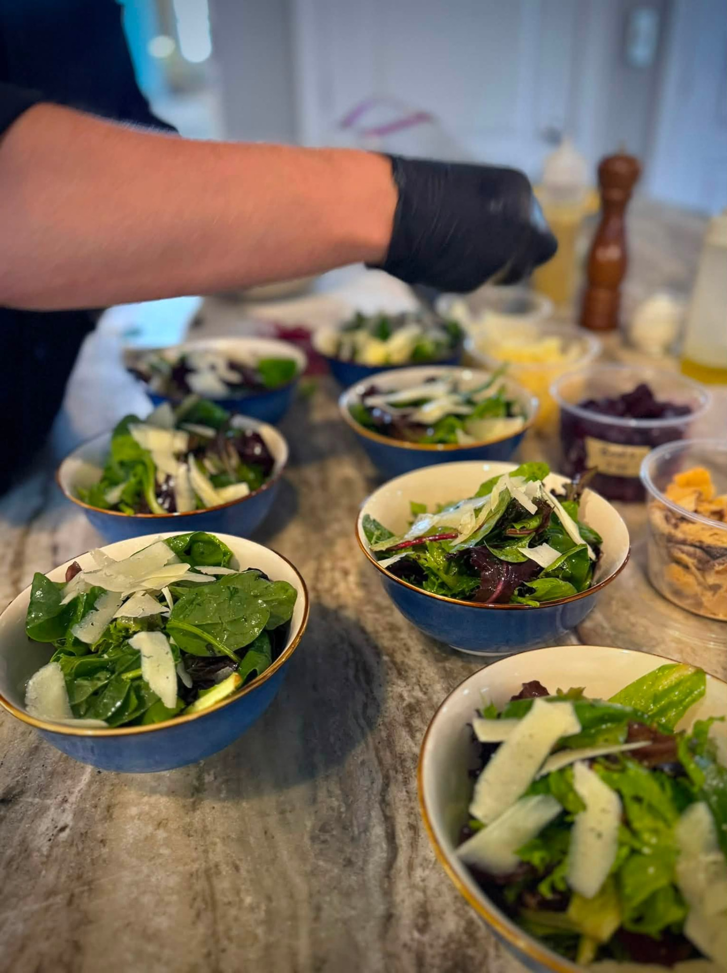 Chef plating individual bowls of mixed greens with shaved parmesan on a marble counter