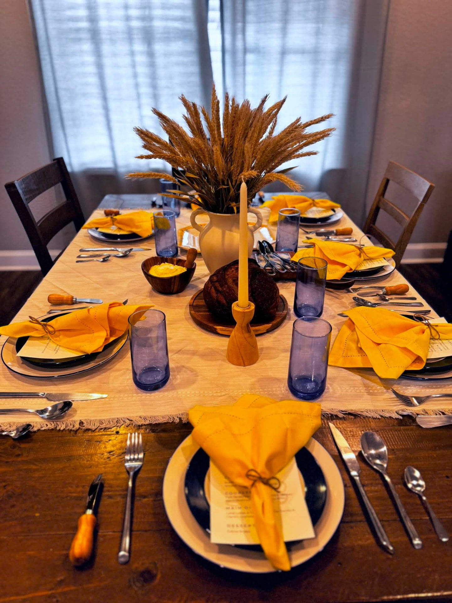 Long dinner table set with printed menus, yellow napkins, blue glassware and a wheat centerpiece